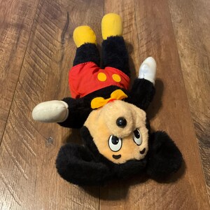 K&ouml;nnte beinhalten: Ein Mickey Mouse Pl&uuml;schtier in einer spielerischen Pose. Das Spielzeug hat einen schwarzen K&ouml;rper, rote Shorts mit gelben Kn&ouml;pfen, gelbe Schuhe und wei&szlig;e Handschuhe. Das Gesicht ist hellbraun mit schwarzen Augen und einer schwarzen Nase.