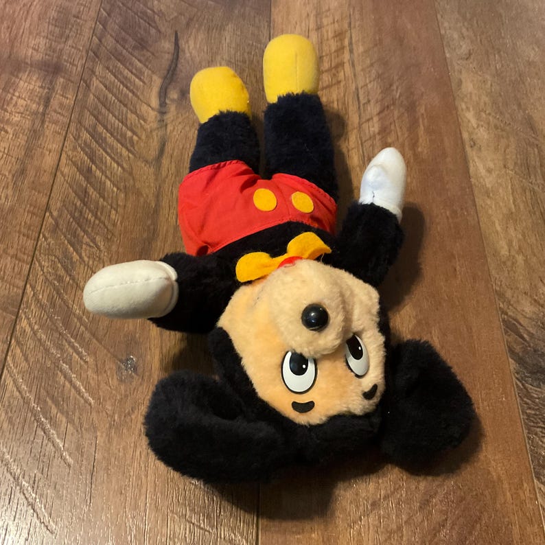 K&ouml;nnte beinhalten: Ein Mickey Mouse Pl&uuml;schtier in einer spielerischen Pose. Das Spielzeug hat einen schwarzen K&ouml;rper, rote Shorts mit gelben Kn&ouml;pfen, gelbe Schuhe und wei&szlig;e Handschuhe. Das Gesicht ist hellbraun mit schwarzen Augen und einer schwarzen Nase.