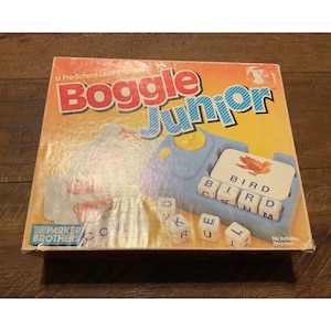 May include: A yellow and blue Boggle Junior game box with a picture of the game board and a bird on a card. The box says "a Pre-School Learning Game" and "Boggle Junior".