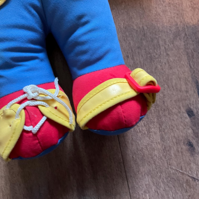 May include: Close-up of a plush doll's legs and feet. The doll's legs are blue, and the feet are red with yellow and red accents. The shoes have yellow laces and a red buckle. The doll is on a wooden surface.
