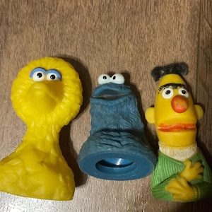 May include: Three Sesame Street character figurines: Big Bird, Cookie Monster, and Bert. Big Bird is yellow, Cookie Monster is blue, and Bert is yellow with a green sweater. A pink measuring tape is next to the figurines.