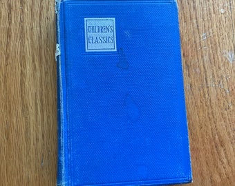 May include: A blue hardcover book titled "CHILDREN'S CLASSICS" in a white rectangular box. The book has a textured cover and is resting on a wooden surface. The book's spine shows signs of wear.