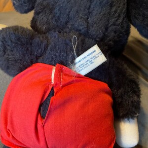 K&ouml;nnte beinhalten: Ein schwarzes Pl&uuml;schtier mit rotem Kleidungsst&uuml;ck. Das Spielzeug hat wei&szlig;e F&uuml;&szlig;e und ein Etikett mit der Aufschrift "WALT DISNEY PRODUCTIONS". Das rote Kleidungsst&uuml;ck hat eine wei&szlig;e Naht.