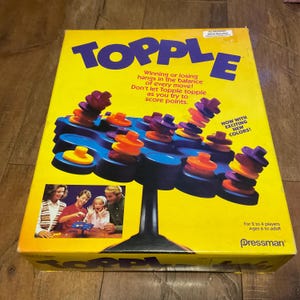 May include: A yellow box for the game Topple, with the game's name in large purple letters. The game features a blue platform with colorful, stacked discs. The box includes text that reads "Winning or losing hangs in the balance of every move!"