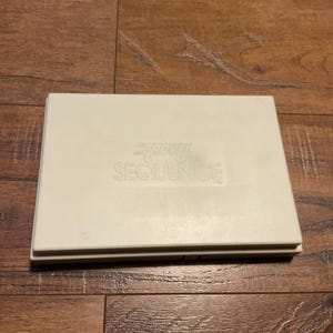 May include: A rectangular, off-white game box with the words "TRAVEL SEQUENCE" printed on the top. The box is closed and appears to be made of plastic. The game is likely a travel-sized version of the popular board game.