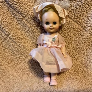 May include: A vintage doll with blonde hair, blue eyes, and a pink dress. She is wearing a pink hat with a white lace trim. The doll is sitting on a brown fuzzy surface.