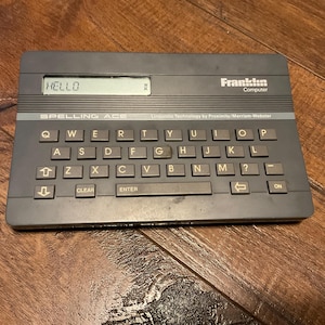 May include: A gray Franklin Spelling Ace electronic dictionary with a keyboard and an LCD screen displaying the word "HELLO". The device has a rectangular shape and features the brand name "Franklin Computer".
