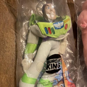 Może przedstawiać: Pluszowa zabawka Buzz Lightyear z serii Toy Story w kolorze zielonym i białym. Zabawka ma na sobie kostium kosmicznego strażnika z logo Burger King na piersi. Zabawka jest zapakowana w przezroczysty plastikowy worek z logo Burger King i tekstem "Ten worek nie jest zabawką. Proszę wyrzucić."