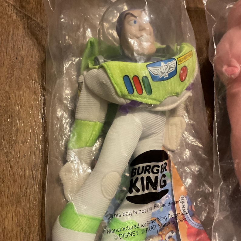 Może przedstawiać: Pluszowa zabawka Buzz Lightyear z serii Toy Story w kolorze zielonym i białym. Zabawka ma na sobie kostium kosmicznego strażnika z logo Burger King na piersi. Zabawka jest zapakowana w przezroczysty plastikowy worek z logo Burger King i tekstem "Ten worek nie jest zabawką. Proszę wyrzucić."