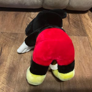 Pu&ograve; includere: Un peluche di Topolino, visto da dietro, con pelliccia nera, pantaloncini rossi, scarpe gialle e guanti bianchi. Il giocattolo &egrave; appoggiato su una superficie di legno.