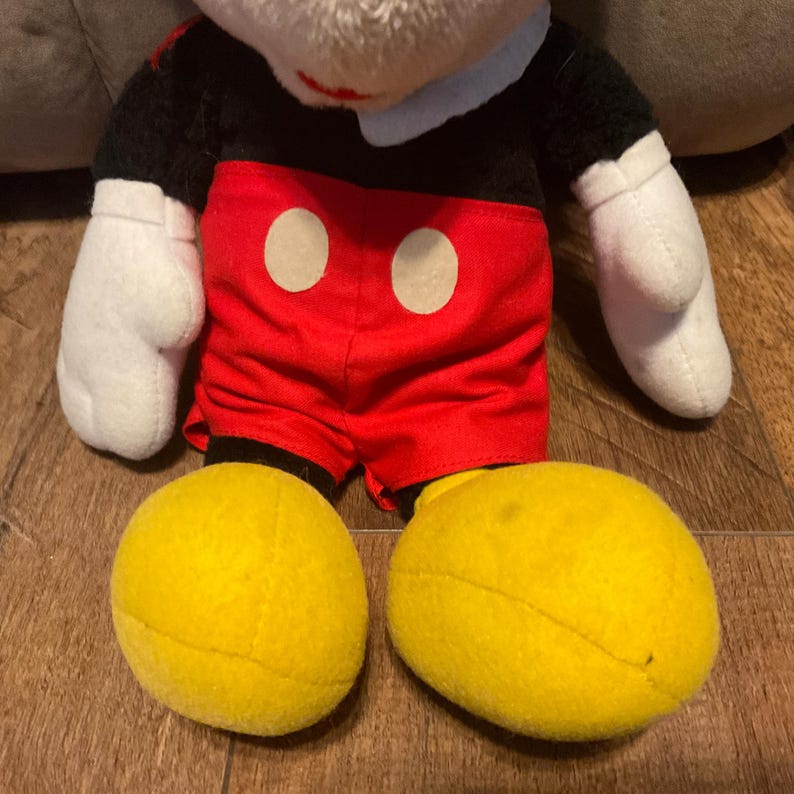 K&ouml;nnte beinhalten: Eine Mickey Mouse Pl&uuml;schpuppe. Die Puppe hat ein wei&szlig;es Gesicht, einen schwarzen K&ouml;rper, rote Shorts mit zwei wei&szlig;en Kreisen, wei&szlig;e Handschuhe und gro&szlig;e gelbe F&uuml;&szlig;e. Eine klassische Disney-Figur.