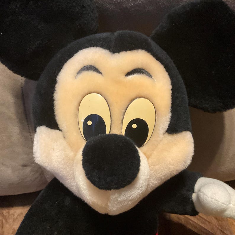 Pu&ograve; includere: Un peluche di Topolino con testa e orecchie nere, viso color crema e grandi occhi gialli. Il giocattolo ha un naso nero e bocca e mani bianche. Il peluche &egrave; un classico personaggio Disney.