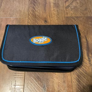 May include: A black zippered case with a blue trim and the word "Boggle" in an orange and blue oval. The case is rectangular and appears to be made of a durable fabric. The background is a wooden surface.