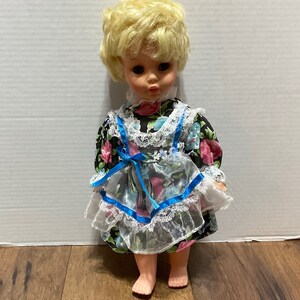 May include: A vintage doll with blonde hair wearing a black and white floral dress with blue trim and white lace. The doll has blue eyes and a sweet expression.