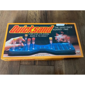 May include: A box for the Quicksand game, a sand race against time game for 2 or 4 players, ages 8 to adult. The box is orange and blue with a picture of a hand playing the game. The game tray is blue and has 48 holes. The box says "Complete" and "The Sand Race Against Time".