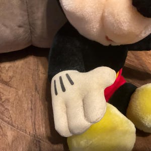 Pu&ograve; includere: Primo piano di un peluche di Topolino. Il giocattolo presenta una testa e un corpo neri, una camicia rossa e scarpe gialle. La mano &egrave; bianca con tre linee nere. Lo sfondo &egrave; una superficie di legno.