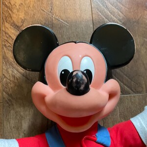 May include: A vintage Mickey Mouse doll with a red shirt, blue overalls, and large black ears. The doll's face is pink with a black nose and white eyes. The doll's arms are white.