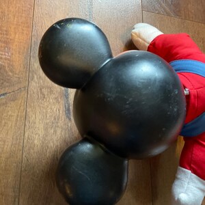 May include: Black Mickey Mouse-shaped bowling ball with three holes. The ball has a glossy finish and is slightly worn. A portion of a red and blue plush toy is visible in the background.