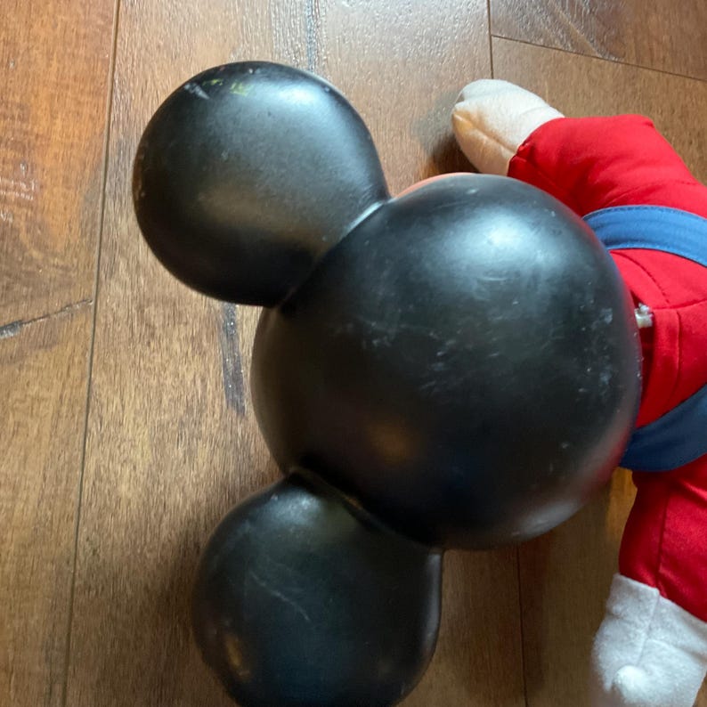 May include: Black Mickey Mouse-shaped bowling ball with three holes. The ball has a glossy finish and is slightly worn. A portion of a red and blue plush toy is visible in the background.