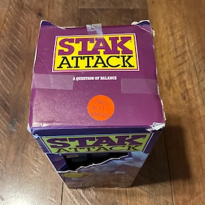 Peut inclure: Bo&icirc;te de jeu Stak Attack violette et jaune. Le titre du jeu est en grandes lettres jaunes avec un contour noir. Le texte "A Question of Balance" est en dessous. Un autocollant orange indique "Has all pieces".