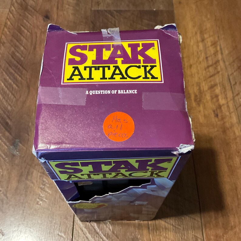 Peut inclure: Bo&icirc;te de jeu Stak Attack violette et jaune. Le titre du jeu est en grandes lettres jaunes avec un contour noir. Le texte "A Question of Balance" est en dessous. Un autocollant orange indique "Has all pieces".