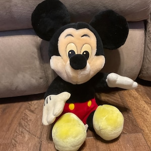 Pu&ograve; includere: Un peluche di Topolino. Il giocattolo presenta un corpo, orecchie e testa neri, con un viso color camoscio, guanti bianchi, pantaloncini rossi e piedi gialli. Il classico personaggio Disney &egrave; seduto su un pavimento di legno.