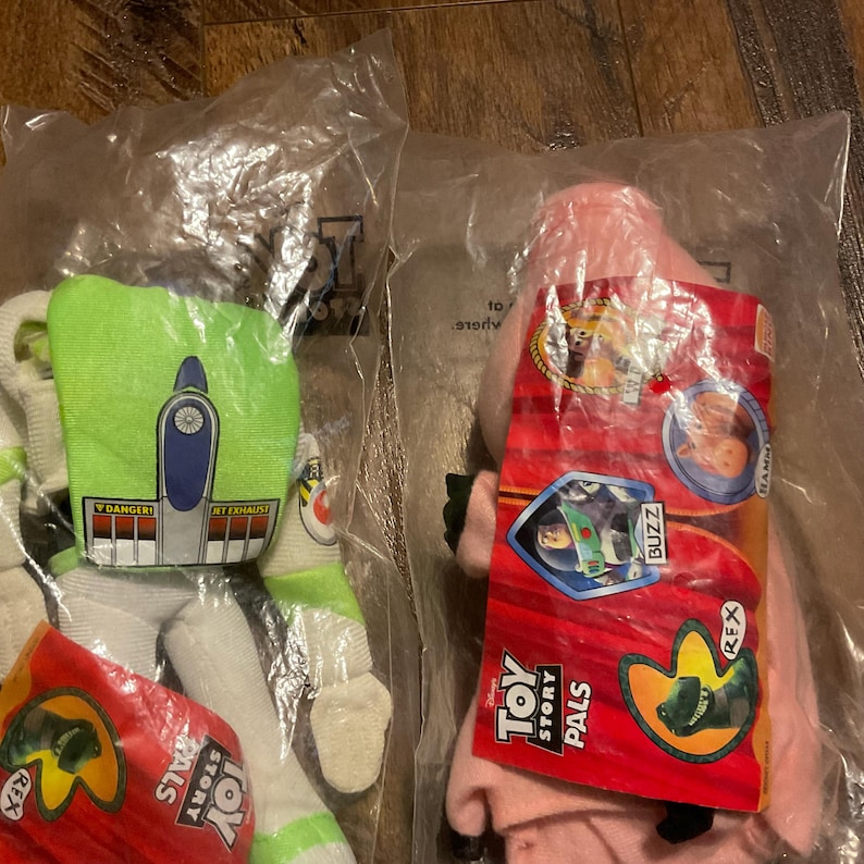 Może przedstawiać: Dwie pluszowe zabawki Toy Story Pals w oryginalnym opakowaniu. Pluszowe zabawki to Buzz Astral i Rex. Buzz Astral jest zielony i biały, a Rex jest r&oacute;żowy. Opakowanie jest czerwone i zawiera zdjęcia postaci.
