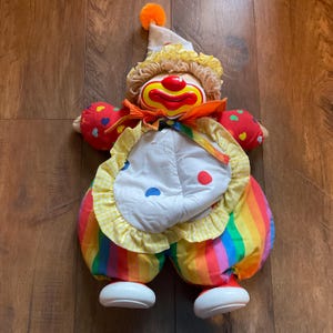 May include: A colorful clown doll with a white bib, rainbow pants, and red heart-shaped hands. The clown has a yellow ruffled collar, a white hat with an orange pom-pom, and a painted face with a red smile.