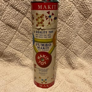 May include: A vintage cardboard tube for a Jumbo Makit toy construction set. The tube is red, white, and blue with illustrations of the toy's parts and the text "A Quality Toy with parts that move and wheels that turn."  "Jumbo Makit 292 Pieces" and "Makit Toy" are also printed on the tube.