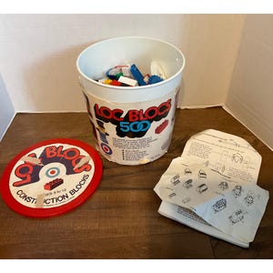 May include: A white plastic bucket filled with colorful plastic building blocks. The bucket has a red and white lid with the words "Loc Blocs Construction Blocks" printed on it. The bucket also has a label that says "Loc Blocs 500".