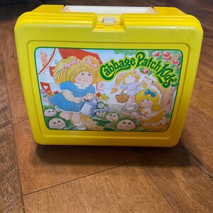 May include: A vintage yellow plastic lunchbox featuring a colorful graphic of Cabbage Patch Kids. The lunchbox has a white handle and clasp. The graphic shows three Cabbage Patch Kids, a house, and the words "Cabbage Patch Kids".