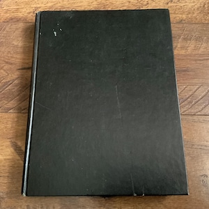 May include: A closed, hardcover book with a black cover. The book is rectangular and appears to be lying on a wooden surface. The cover has some minor wear and tear, with a few small white marks.