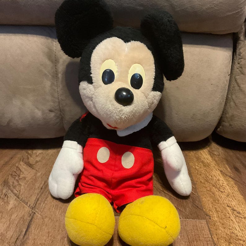 K&ouml;nnte beinhalten: Eine Pl&uuml;schpuppe von Mickey Mouse mit schwarzen Ohren, einer schwarzen Nase und einem cremefarbenen Gesicht. Die Puppe tr&auml;gt eine rote Hose mit zwei wei&szlig;en Kreisen und wei&szlig;en Handschuhen. Zwei gelbe Tennisb&auml;lle liegen vor der Puppe.