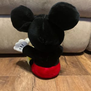 Pu&ograve; includere: Un peluche di Topolino, visto da dietro, con grandi orecchie nere e un corpo nero. Il giocattolo ha un fondo rosso, pantaloncini gialli e guanti bianchi con dettagli neri. Il giocattolo &egrave; appoggiato su una superficie di legno.