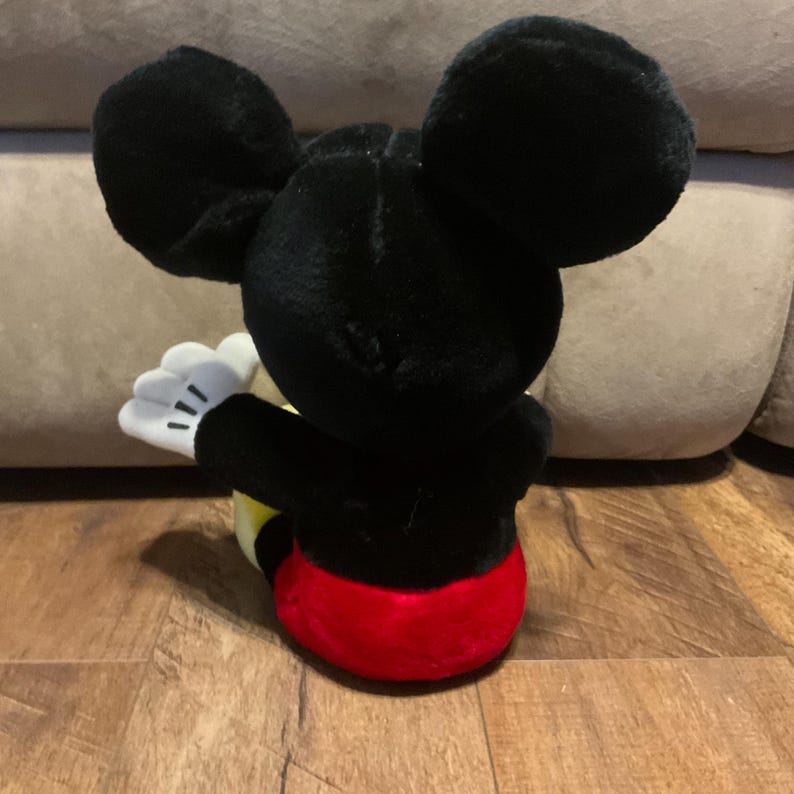 Pu&ograve; includere: Un peluche di Topolino, visto da dietro, con grandi orecchie nere e un corpo nero. Il giocattolo ha un fondo rosso, pantaloncini gialli e guanti bianchi con dettagli neri. Il giocattolo &egrave; appoggiato su una superficie di legno.