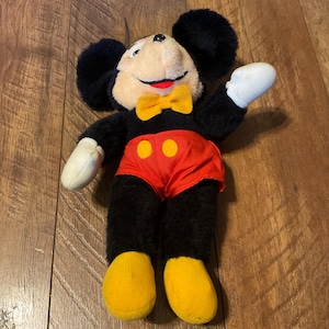 K&ouml;nnte beinhalten: Ein Vintage-Mickey-Mouse-Pl&uuml;schtier. Das Pl&uuml;schtier hat schwarzes Fell, ein cremefarbenes Gesicht und eine gelbe Fliege. Mickey tr&auml;gt rote Shorts mit gelben Kn&ouml;pfen und gelbe Schuhe. Das Spielzeug liegt auf einer Holzoberfl&auml;che.