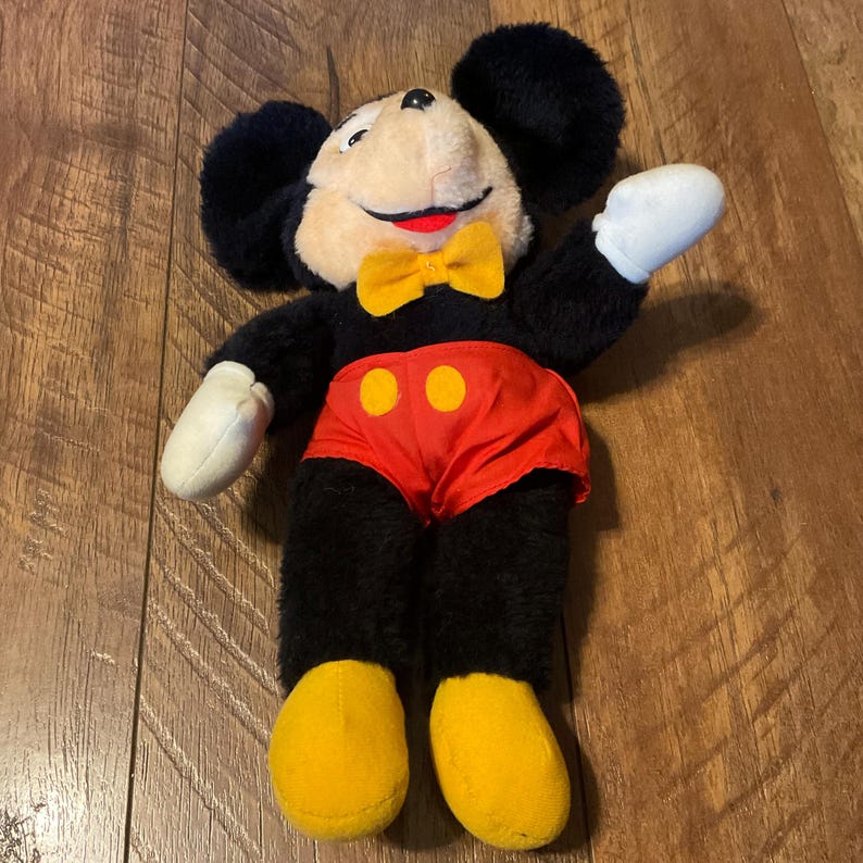 K&ouml;nnte beinhalten: Ein Vintage-Mickey-Mouse-Pl&uuml;schtier. Das Pl&uuml;schtier hat schwarzes Fell, ein cremefarbenes Gesicht und eine gelbe Fliege. Mickey tr&auml;gt rote Shorts mit gelben Kn&ouml;pfen und gelbe Schuhe. Das Spielzeug liegt auf einer Holzoberfl&auml;che.