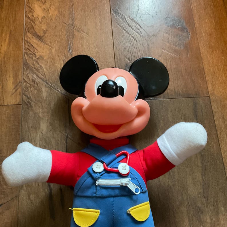 May include: A Mickey Mouse plush doll. It features a red top, blue dungarees, and white gloves. The doll has a large, smiling face with black ears and a black nose. The dungarees have yellow pockets and a zipper.