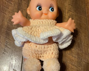Könnte beinhalten: Eine Vintage-Babypuppe mit pfirsichfarbenem Gesicht, blauen Augen und einem gehäkelten Outfit. Das Outfit der Puppe umfasst ein pfirsichfarbenes Oberteil mit weißem Rüschenbesatz, passende Shorts und flauschige pfirsichfarbene Pompons an den Füßen. Die Puppe befindet sich auf einer Holzoberfläche.