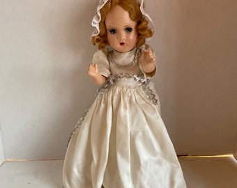 May include: A vintage doll with auburn hair and blue eyes, wearing a cream-colored satin dress with silver trim. The doll has a lace bonnet and is posed with its arms outstretched. A classic collectible toy.