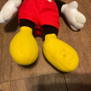 K&ouml;nnte beinhalten: Eine Mickey Mouse-Pl&uuml;schpuppe mit schwarzem Fell, roter Hose, gelben F&uuml;&szlig;en und wei&szlig;en Handschuhen. Die F&uuml;&szlig;e der Puppe sind gro&szlig; und abgerundet, mit einem kleinen Fleck auf einem Fu&szlig;. Die Puppe liegt auf einer Holzoberfl&auml;che.