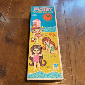 May include: A box of Flatsy paper dolls with a beach theme. The box features three girls with long hair wearing swimsuits. The text on the box reads "Flatsy Paper Dolls" and "No Scissors Necessary".
