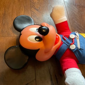 May include: A vintage Mickey Mouse plush toy. The toy has a large black head with a pink face, black eyes, and a black nose. Mickey wears a red shirt, blue dungarees, and white gloves. The toy is lying on a wooden surface.
