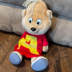 May include: A plush toy of Alvin from the cartoon series, The Chipmunks. The doll has a tan face, blonde hair, and is wearing a red shirt with a yellow "A" on it. The doll has blue and white shoes.