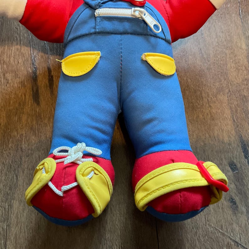 May include: A plush doll with a red top, blue dungarees, and red shoes. The dungarees have yellow pocket details and a zip. The shoes have yellow accents and white laces. The doll is on a wooden surface.