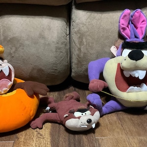 May include: Three plush toys of the cartoon character Taz from Looney Tunes. One is wearing a purple costume with bunny ears, one is wearing a pumpkin costume, and one is brown and has a mischievous expression.
