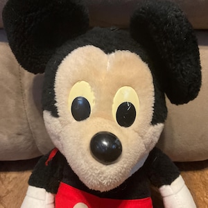 K&ouml;nnte beinhalten: Ein Stofftier von Mickey Mouse mit schwarzen Ohren, einer schwarzen Nase und gro&szlig;en schwarzen Augen. Das Gesicht ist cremefarben, mit gelben Augenakzenten. Das Spielzeug hat ein rotes Hemd mit wei&szlig;en Kn&ouml;pfen und schwarzen &Auml;rmeln. Ein klassischer Disney-Charakter.