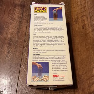 Peut inclure: Bo&icirc;te de jeu Stak Attack vintage. La bo&icirc;te pr&eacute;sente le logo et les instructions du jeu. Le jeu consiste &agrave; retirer des blocs de bois d'une tour sans la faire basculer. La bo&icirc;te est violette et blanche.