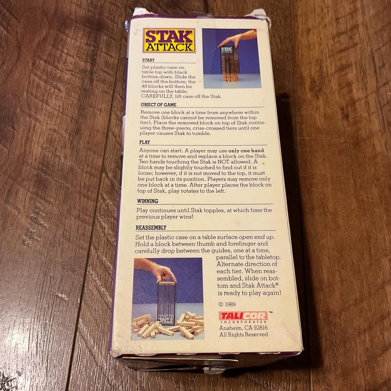 Peut inclure: Bo&icirc;te de jeu Stak Attack vintage. La bo&icirc;te pr&eacute;sente le logo et les instructions du jeu. Le jeu consiste &agrave; retirer des blocs de bois d'une tour sans la faire basculer. La bo&icirc;te est violette et blanche.