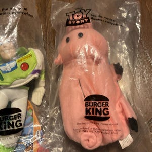 Może przedstawiać: R&oacute;żowa pluszowa zabawka w kształcie świnki z logo Burger King na brzuchu. Zabawka jest zapakowana w przezroczysty plastikowy worek z logo Toy Story i tekstem "Zobacz film w kinach w całej Polsce."